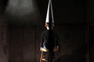 Boy wearing dunce cap sits in dark room.