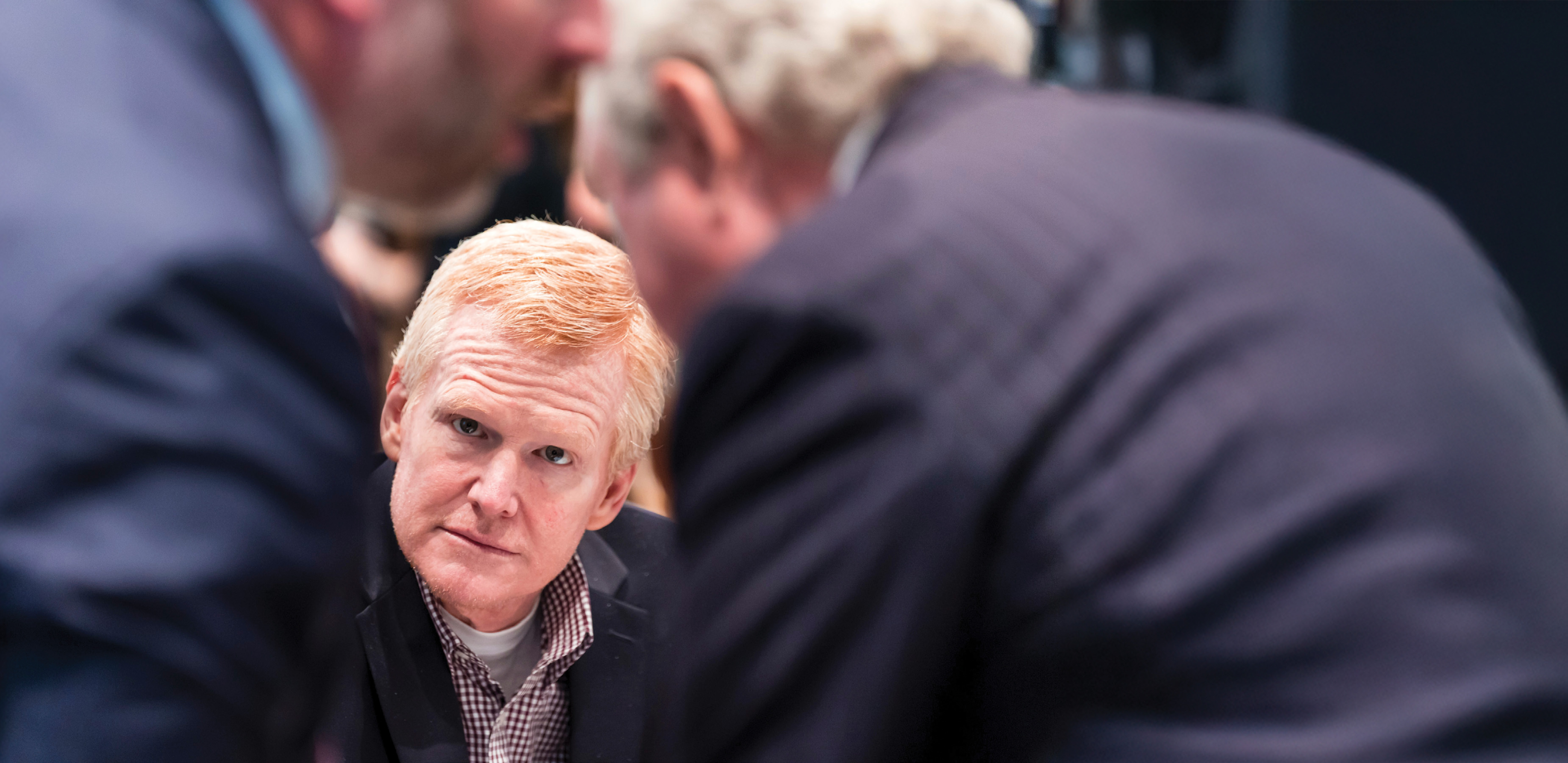 Red haired man seated with two men in suit in the foreground