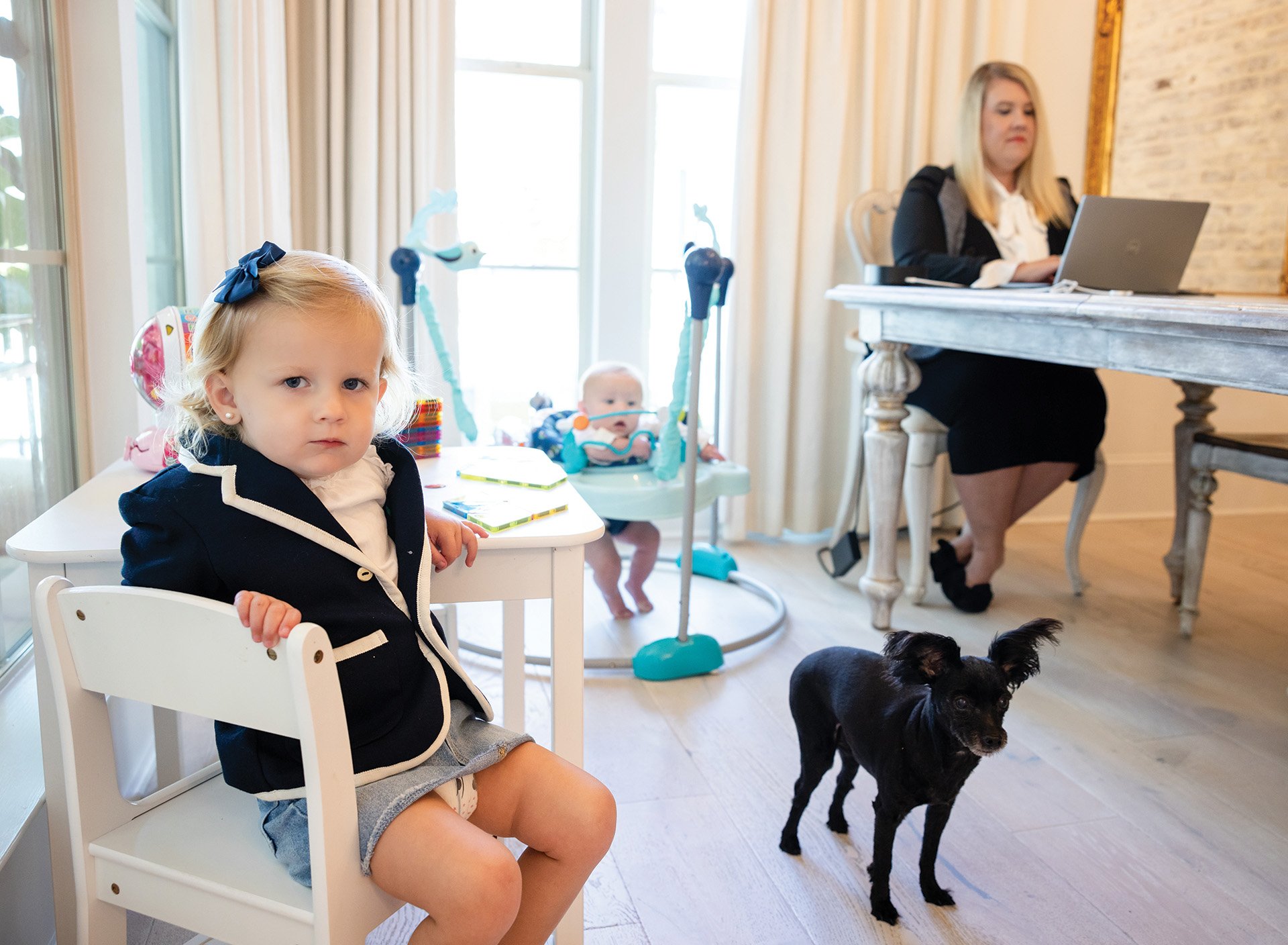 A toddler and dog play in the foreground as a mother works on her laptop in the background