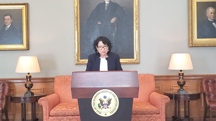 Sonia Sotomayor stands at a podium in front of oil paintings of Supreme Court justices